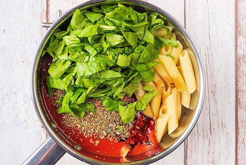 A saucepan containing cooked pasta, chopped spinach, passata and herbs