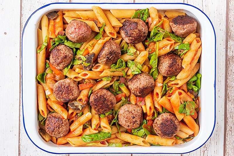 A large rectangular baking dish containing cooked pasta and chopped vegetables all in a tomato sauce. Meatballs have been placed on top
