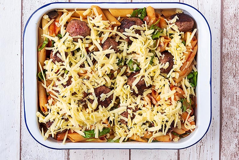 A large rectangular baking dish containing cooked pasta and chopped vegetables all in a tomato sauce. Meatballs have been added and grated cheese sprinkled over everything