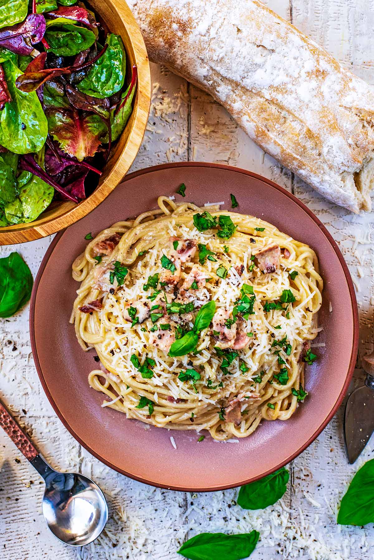 Pasta carbonara on a pink plate. A bowl of salad and some bread are next to it.