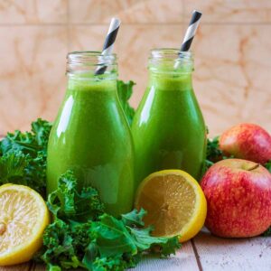 Two small milk bottles of kale smoothie amongst a pile of raw kale leaves.