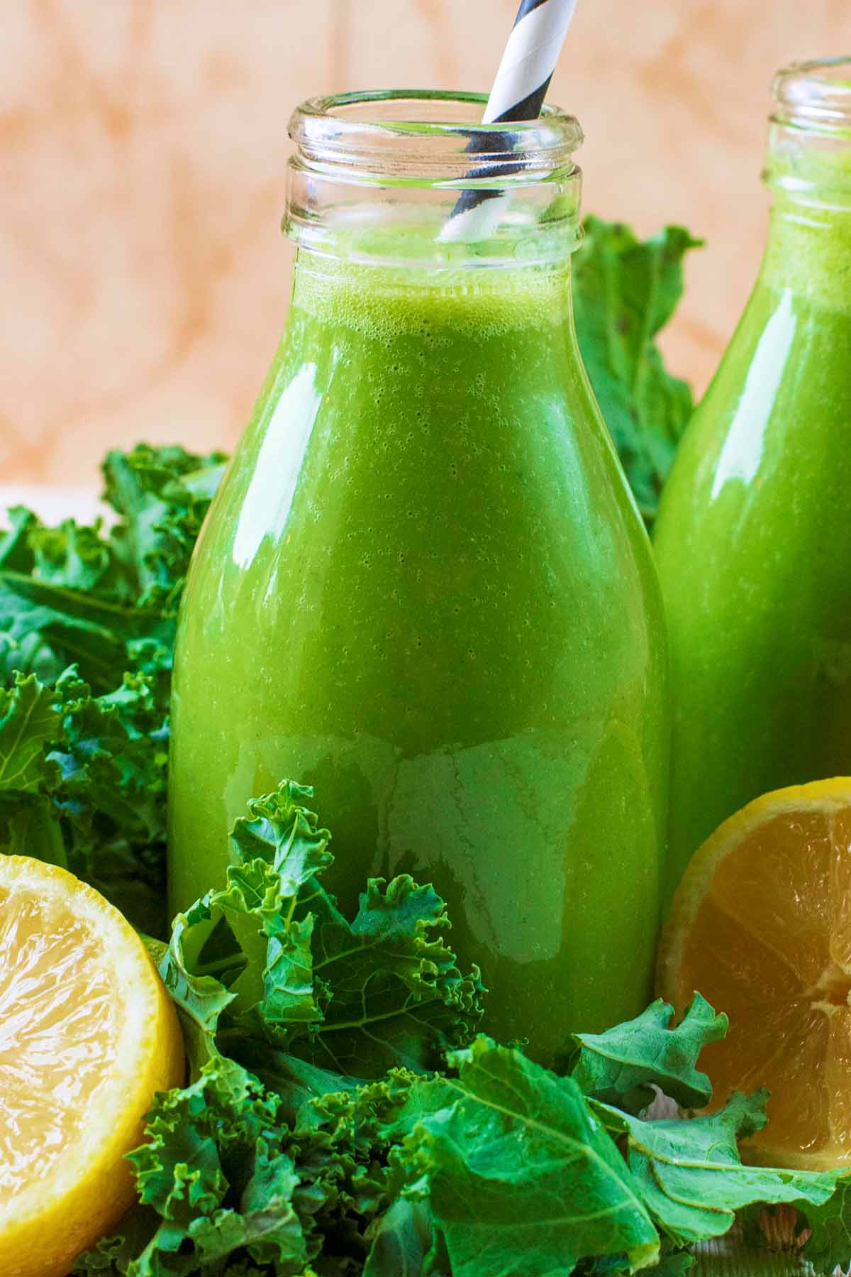 A small glass bottle containing a green smoothie. Green leaves and fruit are surrounding it.