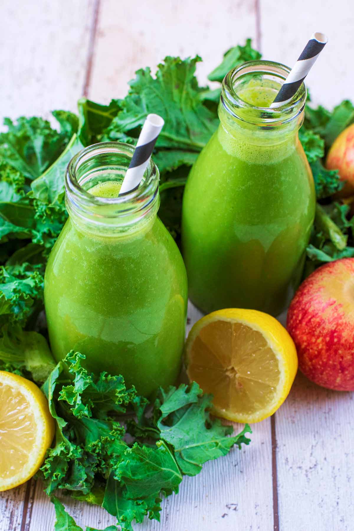 Two bottles of green smoothie surrounded by raw kale, lemon halves and an apple.