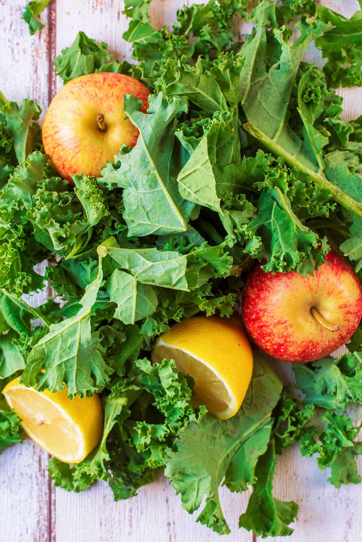 A large pile of raw kale leaves, two apples and two lemon halves all on a wooden surface