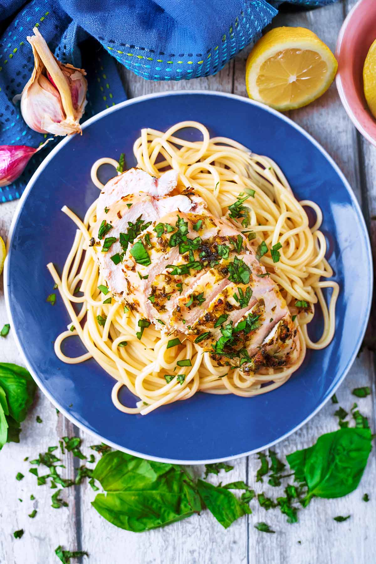 Sliced chicken breast on a bed of spaghetti. A lemon half and some garlic cloves are next to it.