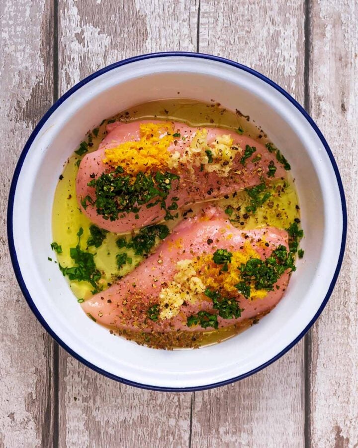 Two raw chicken breasts in a round dish with, lemon juice, zest, oil and chopped basil.