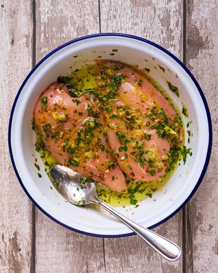 Two raw chicken breasts in a round dish with, lemon juice, zest, oil and chopped basil all mixed together.
