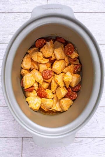 A slow cooker pot containing cooked chicken chunks and sliced chorizo.
