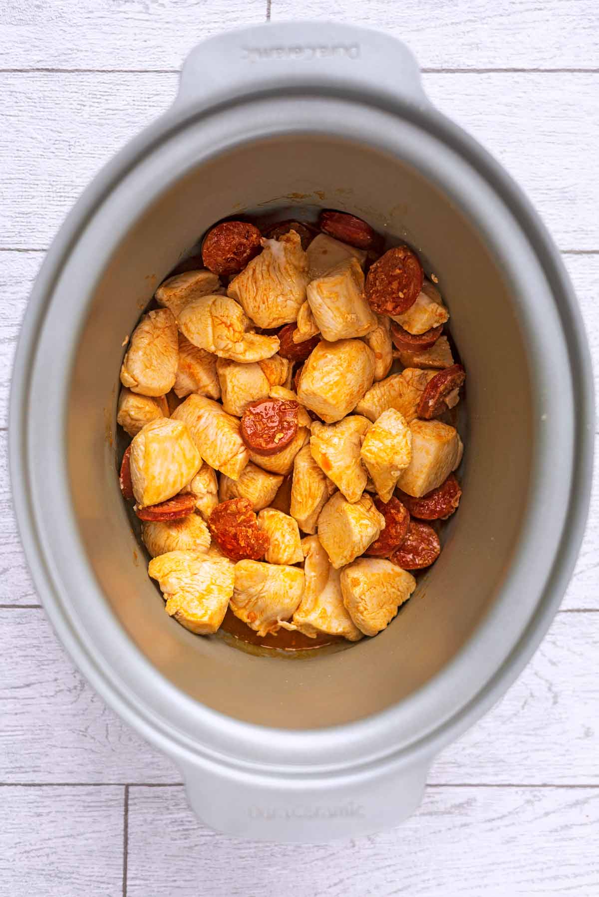A slow cooker pot containing cooked chicken chunks and sliced chorizo.