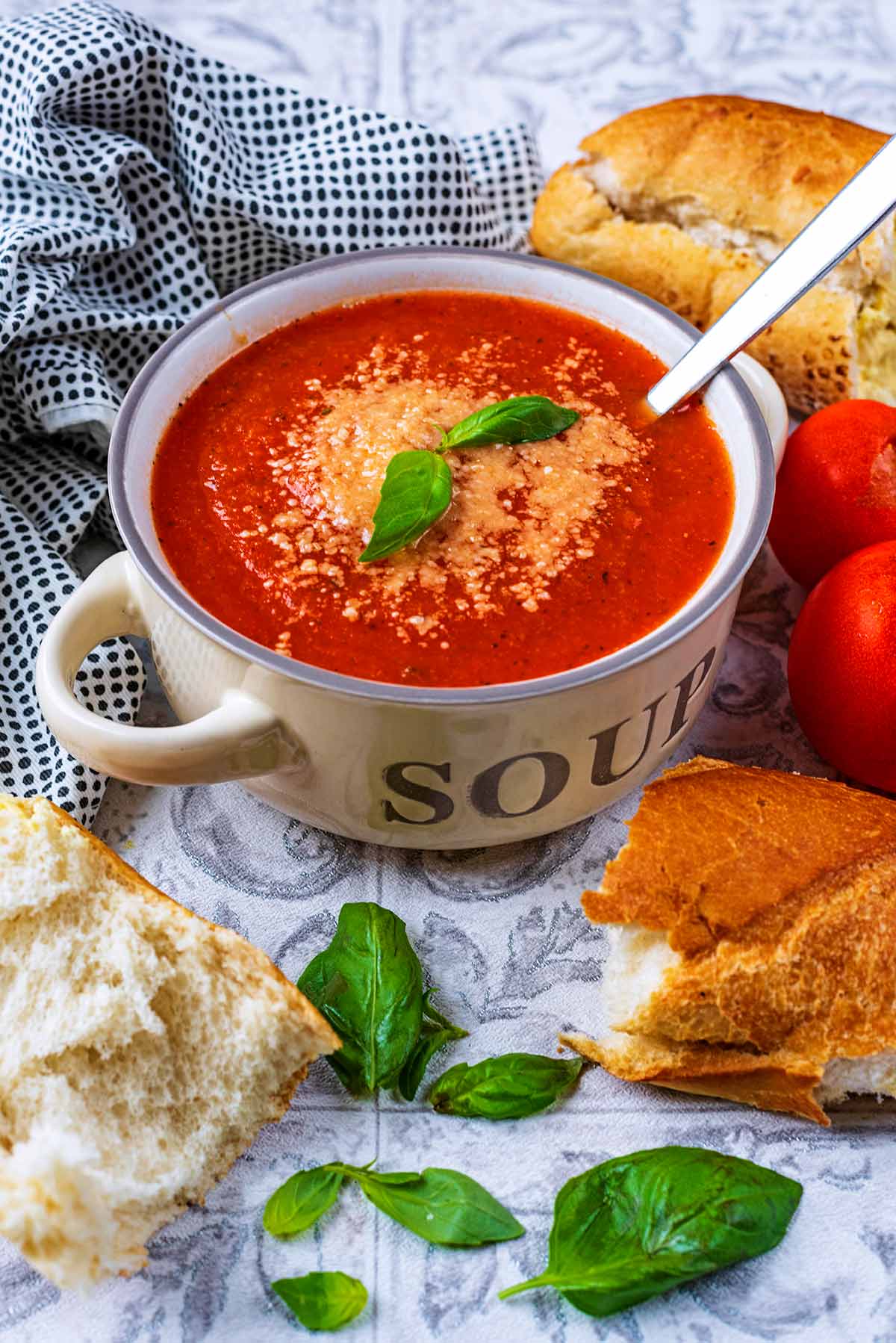 A bowl of tomato soup in a cream soup bowl. Bread and basil leaves are scattered around.