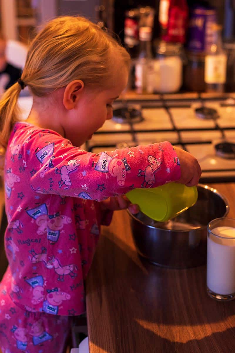 A toddler stood at a work surface pouring from a bowl into a saucepan.