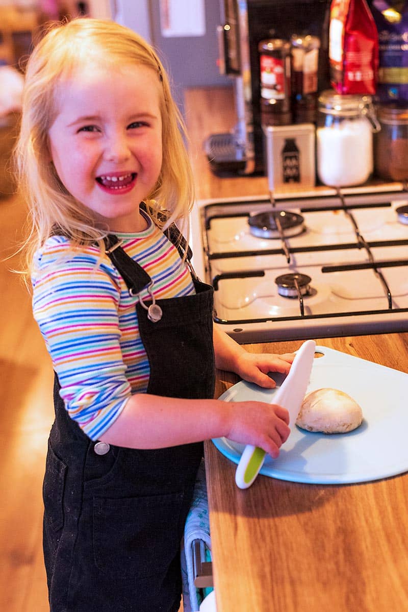 A smiling toddler stood at a work surface holding a plastic knife over a chopping board.