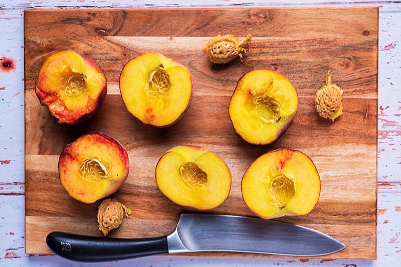 A wooden chopping board with three peaches cut in half. A large chef's knife sits on the board too