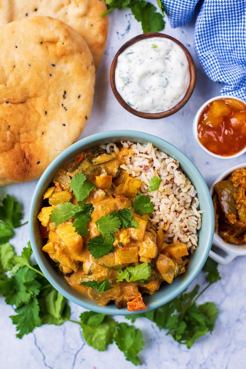 Slow Cooker Vegetable curry in a blue bowl. Naan breads, raita, pickles and chutney sit next to it.