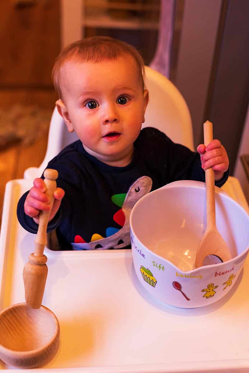 A baby sat in a high chair with a mixing bowl, wooden spoon and rolling pin.