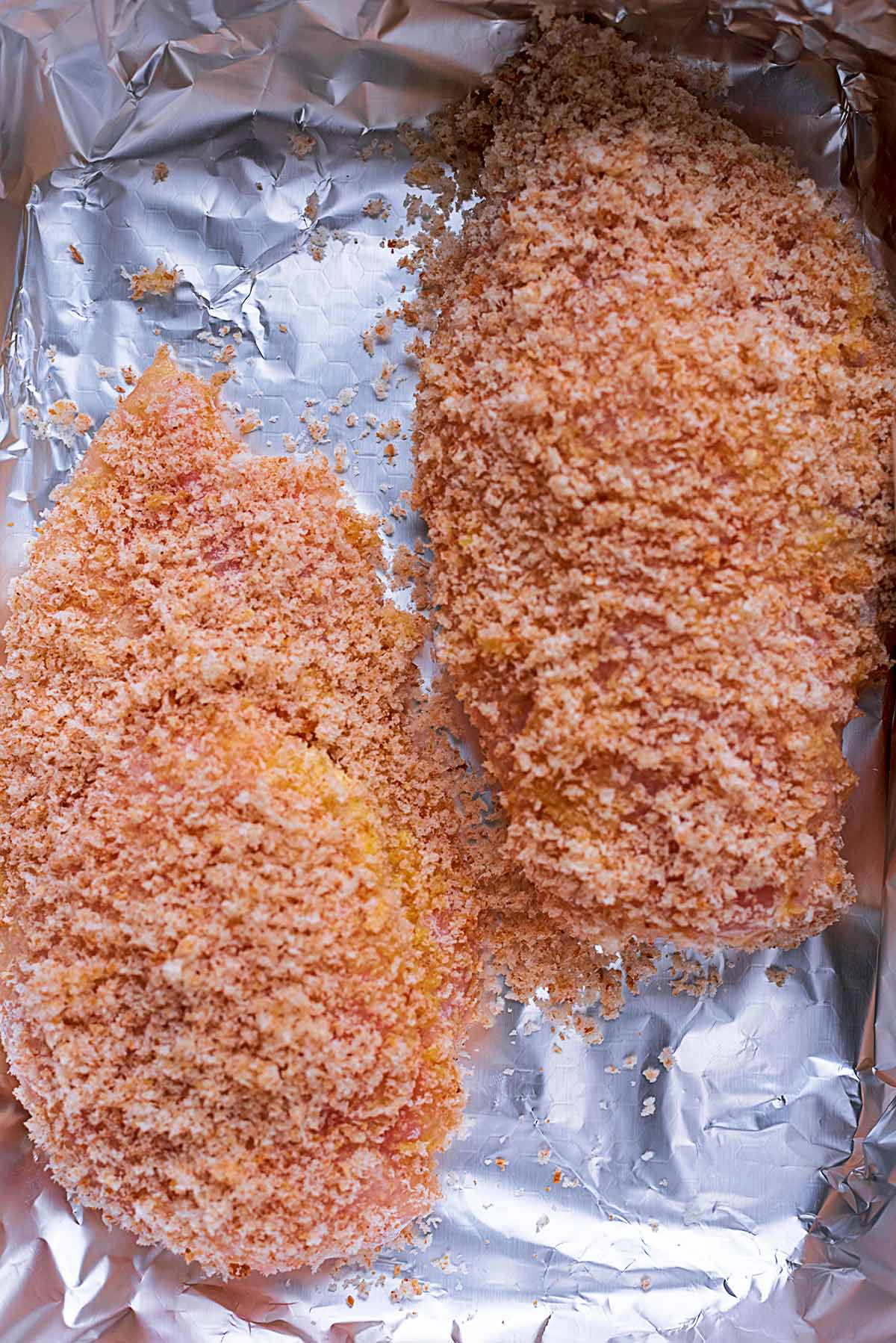 Two chicken breasts coated in bread crumbs on a baking sheet.