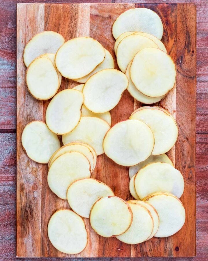 A wooden chopping board with very thinly sliced potatoes.