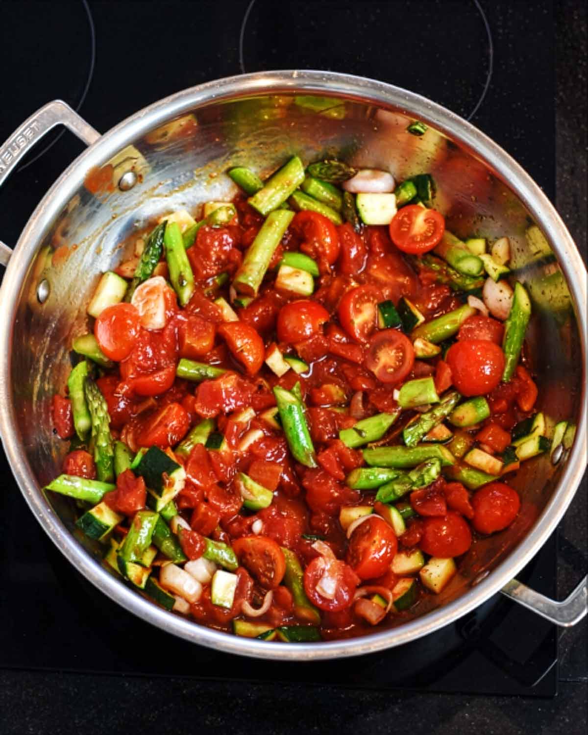 Chopped vegetables and chopped tomatoes cooking in a large silver pan.