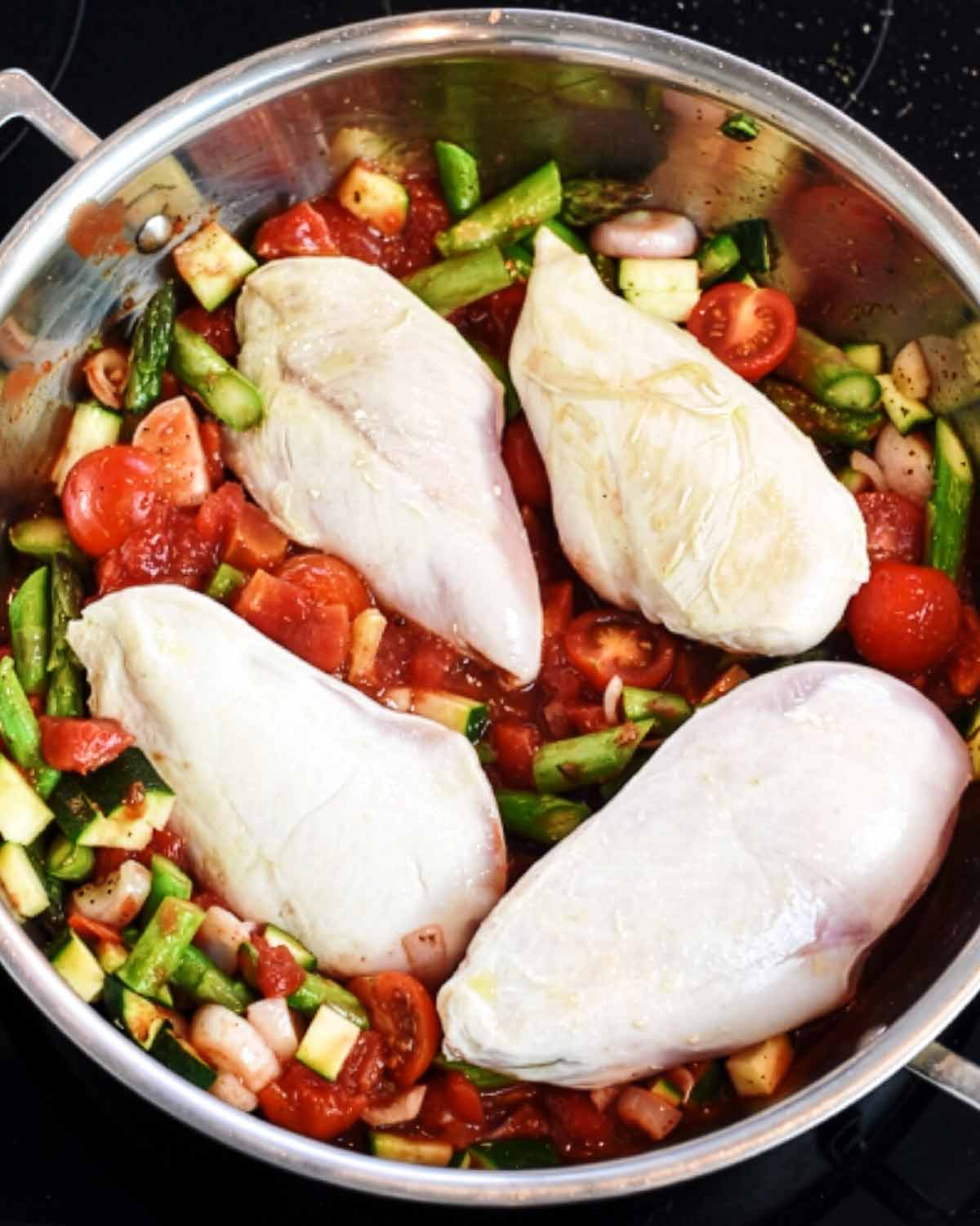 Four part cooked chicken breasts on top of cooked vegetables in a large silver pan.