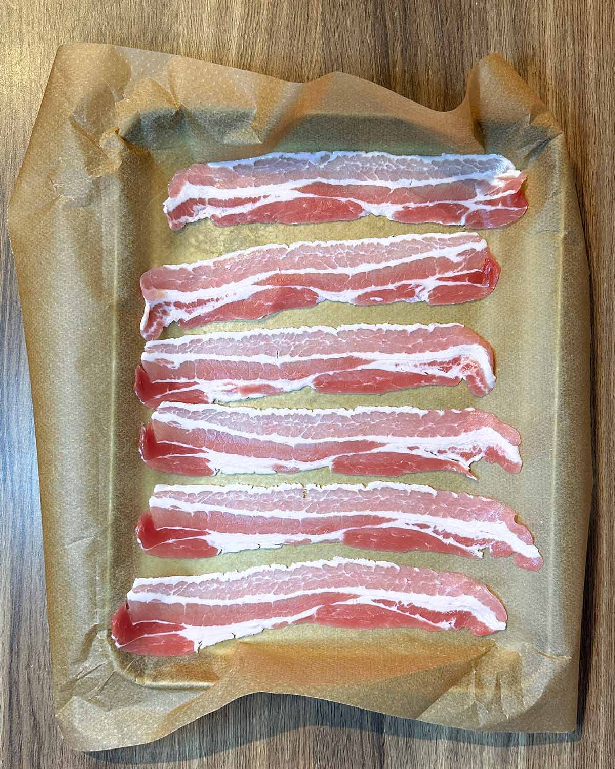 Six rashers of uncooked bacon on a lined baking tray.