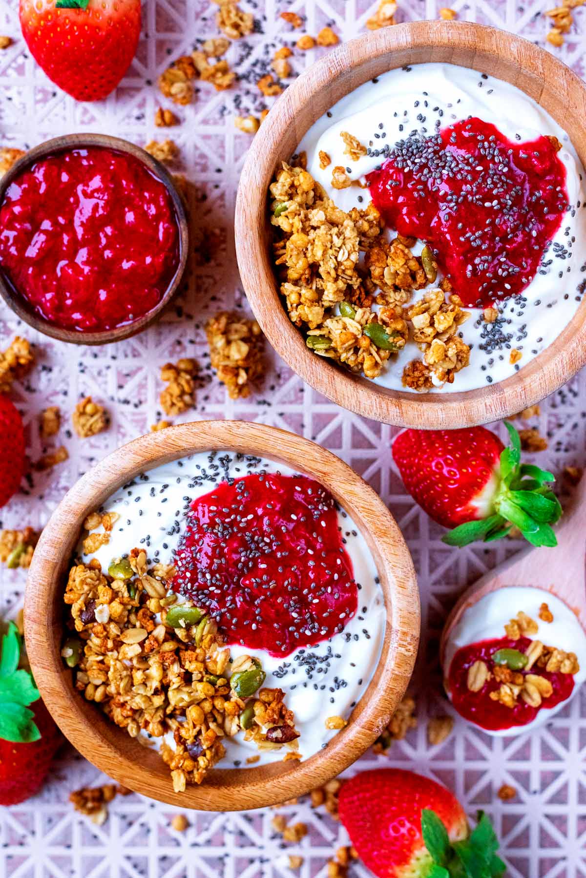 To bowls of yoghurt and granola with strawberry puree on top. Fresh strawberries surround the bowls.