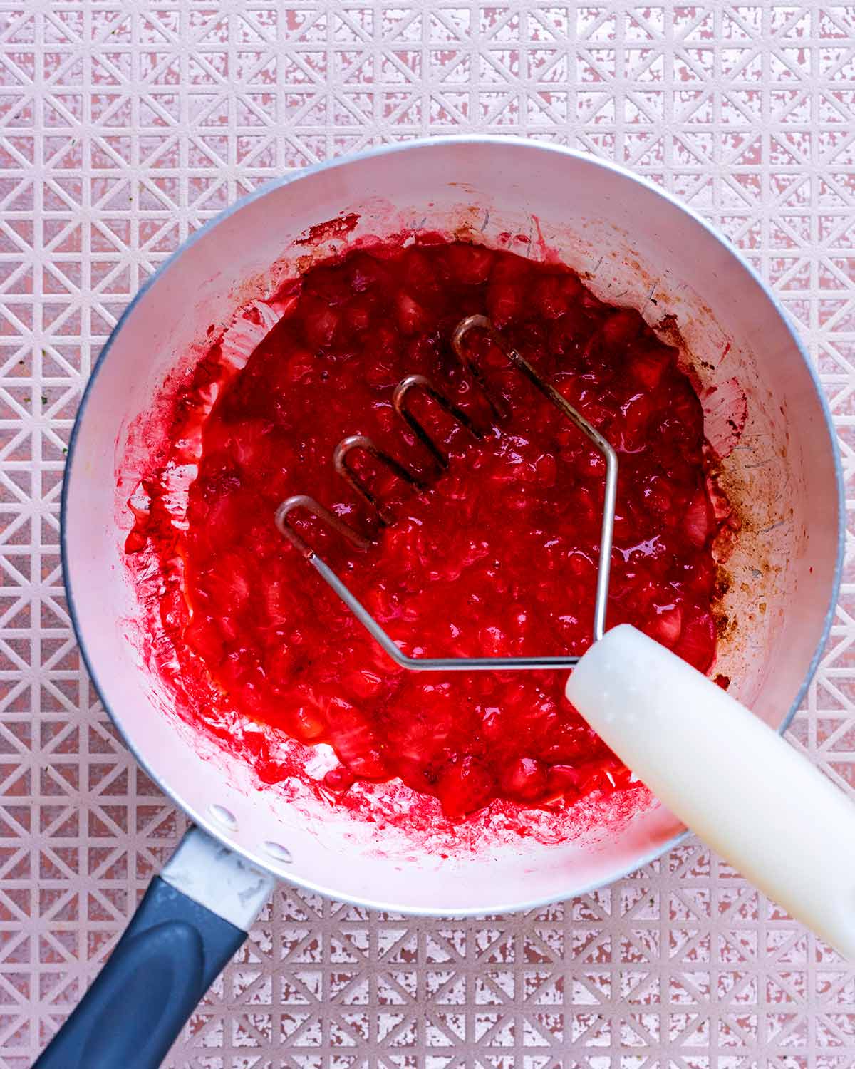 A saucepan with mashed strawberries and a potato masher.