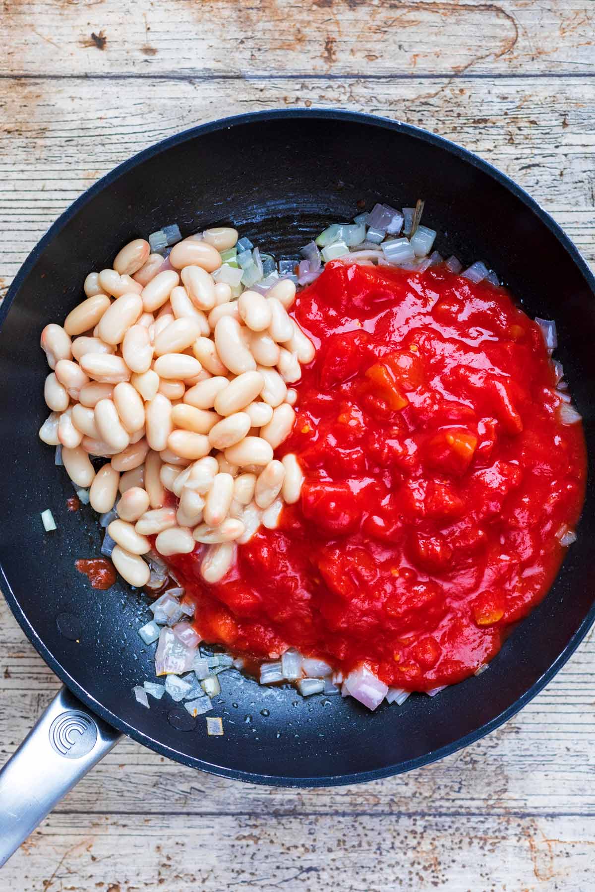 A frying pan with cooked chopped shallots, chopped tomatoes and cannellini beans.