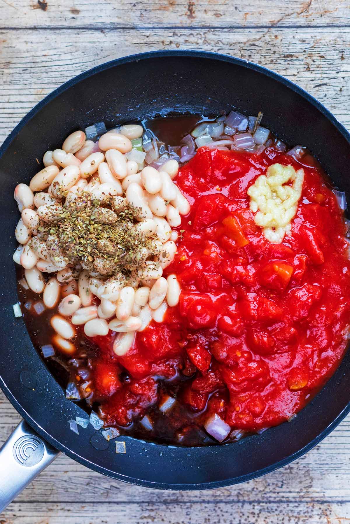 A frying pan with shallots, tomatoes, beans, garlic and herbs.