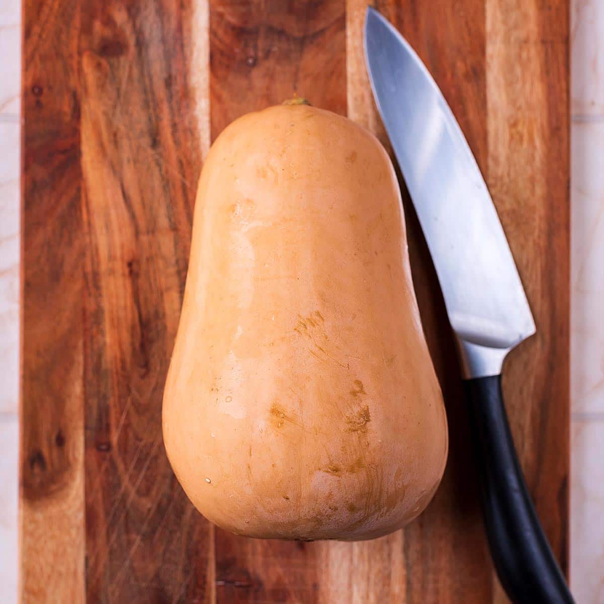 A whole butternut squash and a chef's knife on a wooden chopping board.