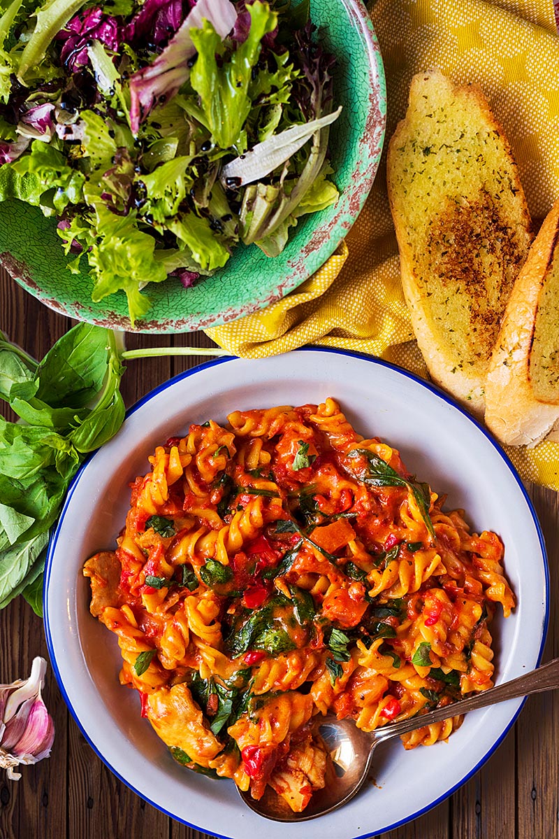 Chicken pasta on a white plate next to garlic bread and a bowl of salad.