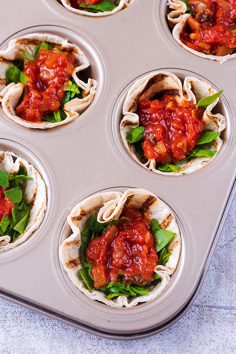 A muffin tin filled with mini tortillas. spinach and marinara.