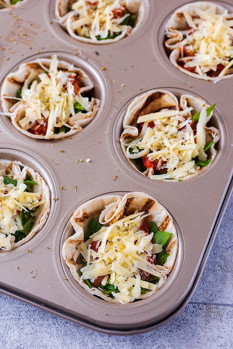A muffin tin filled with mini tortillas. spinach and marinara and topped with grated cheese.