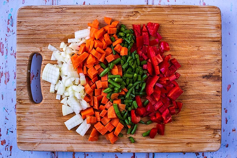 A wooden chopping board with chopped onion, carrot, green beans and red bell pepper.