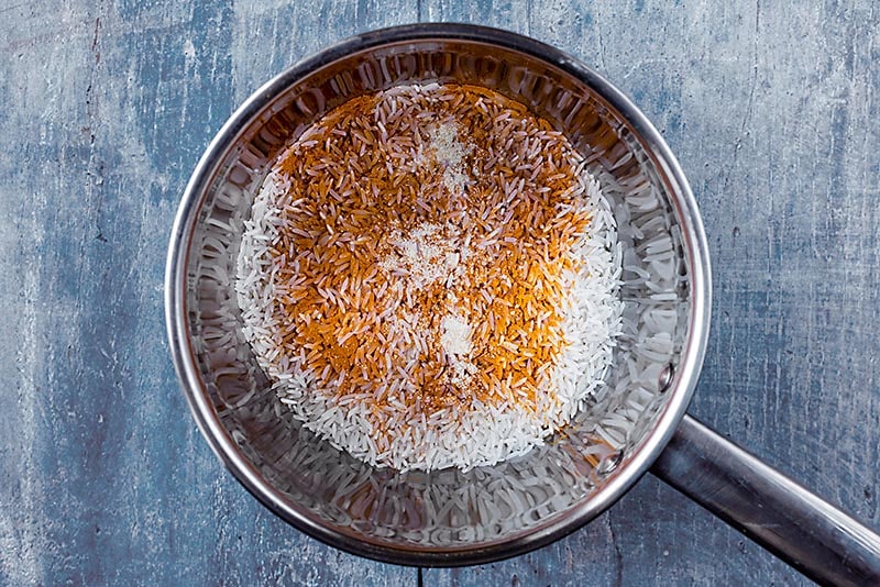 A stainless steel saucepan with uncooked rice and ground turmeric in it.