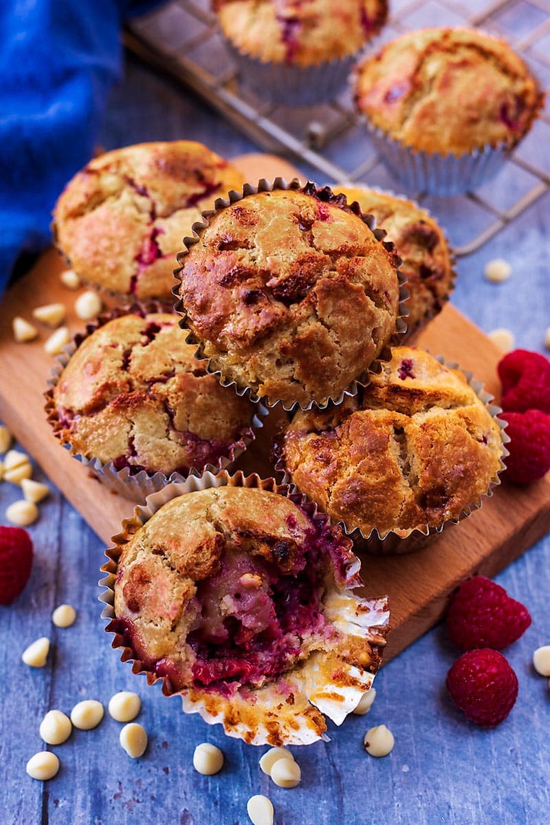 Six raspberry muffins on a board surrounded by raspberries and white chocolate chips