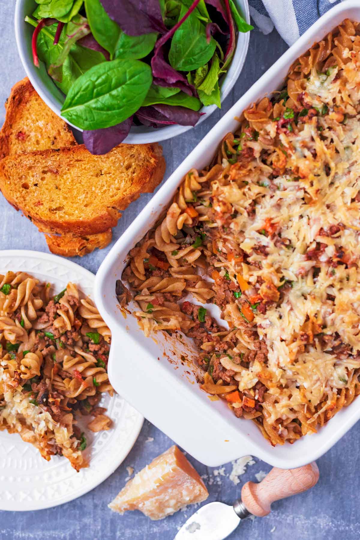 Bolognese pasta in a white baking dish next to a bowl of salad and some garlic bread.