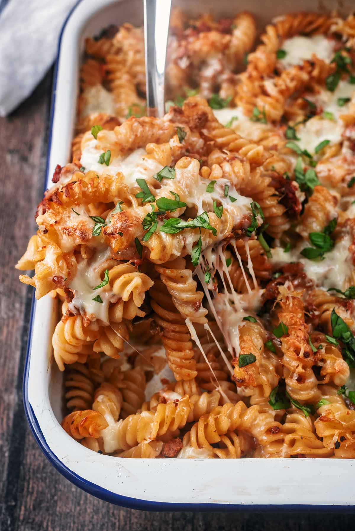 A spoon lifting a portion of pasta bake out of the pan. The melted cheese is stretching.