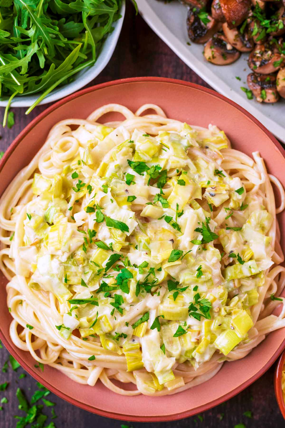 Creamy Leek Pasta in a pink bowl next to a bowl of salad and a dish of roasted mushrooms.