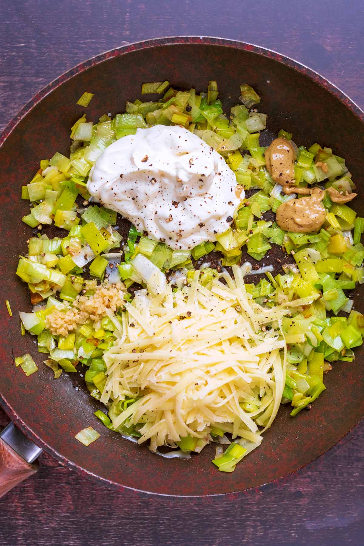 A frying pan with cooked leeks, a dollop of creme fraiche, grated cheese and some mustard.