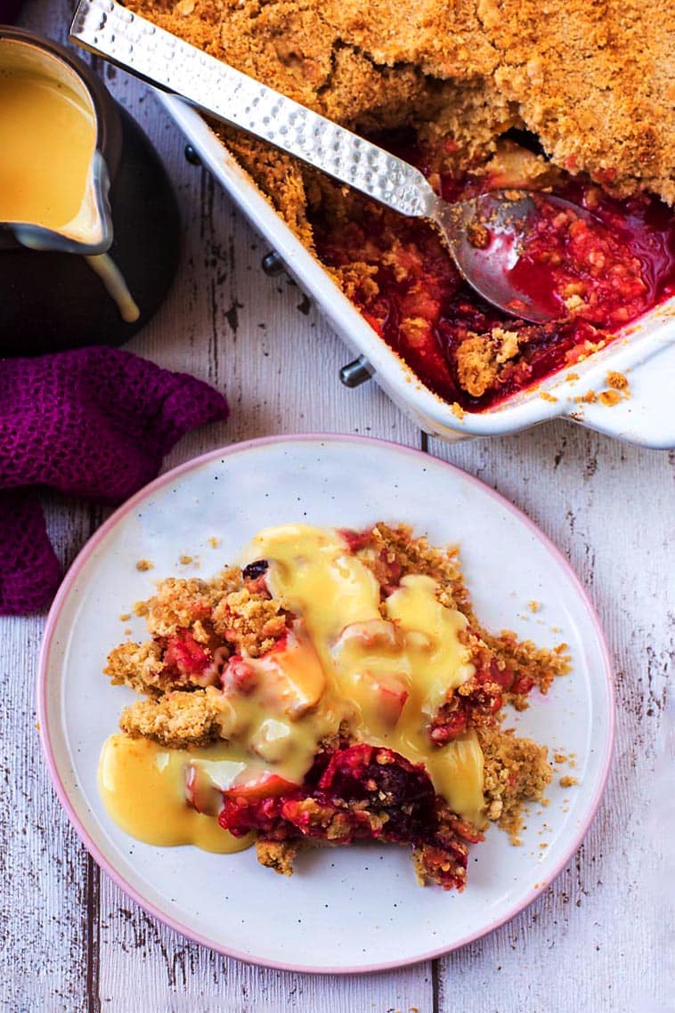 Plum crumble and custard on a plate next to a dish full of crumble.