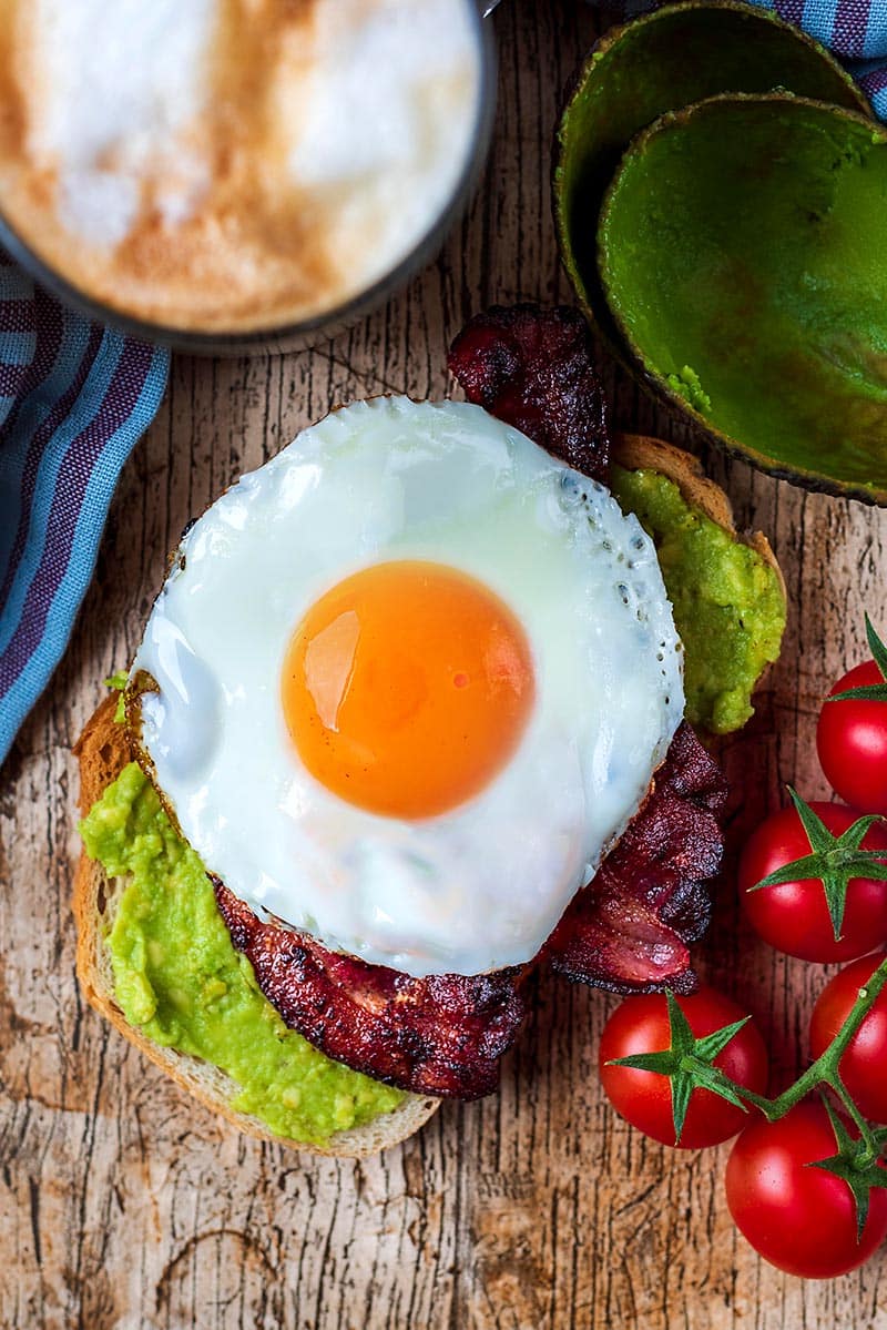 Smashed avocado on toast topped with bacon and a fried egg.