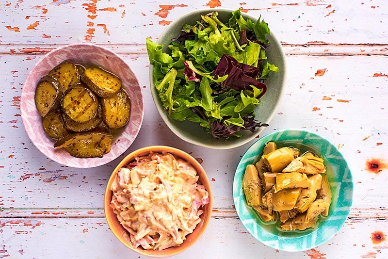 A bowl of salad leaves, a bowl of coleslaw and dishes of sliced zucchini and artichokes