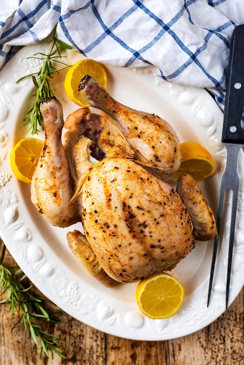 A whole cooked chicken on a large platter with lemon halves and rosemary sprigs.