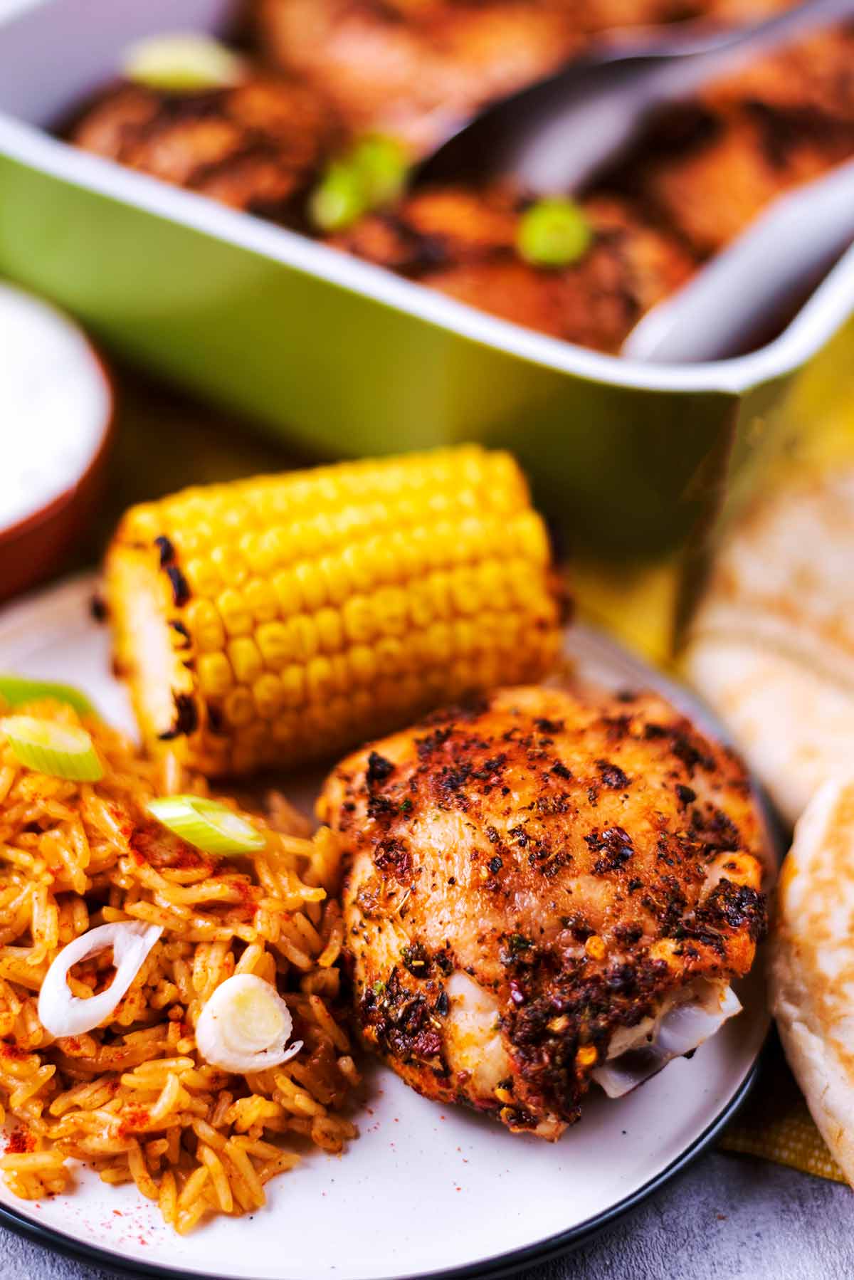 A chicken thigh, corn cob and rice on a plate in front of a baking dish full of chicken.