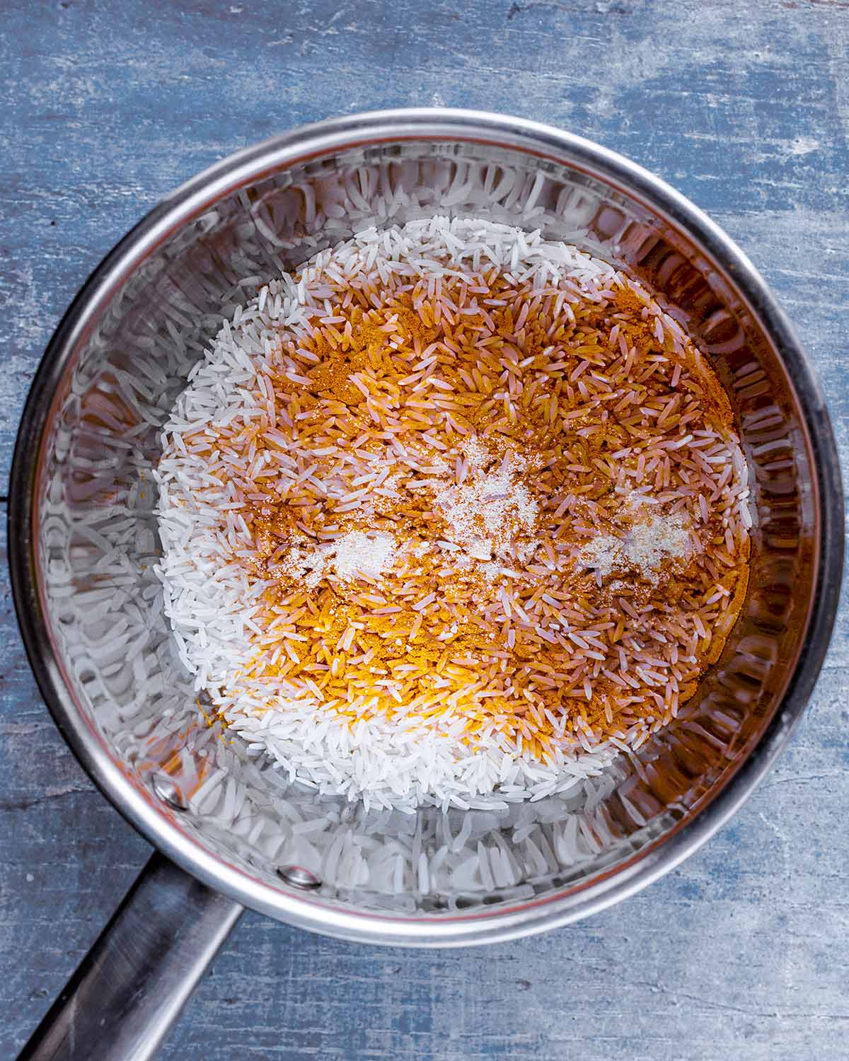 A stainless steel saucepan with uncooked rice and ground turmeric in it.