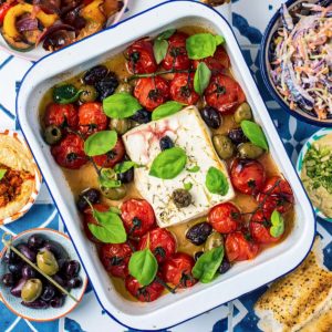Baked Feta with tomatoes in a white baking dish. Olives, hummus, slaw and bread surround it.