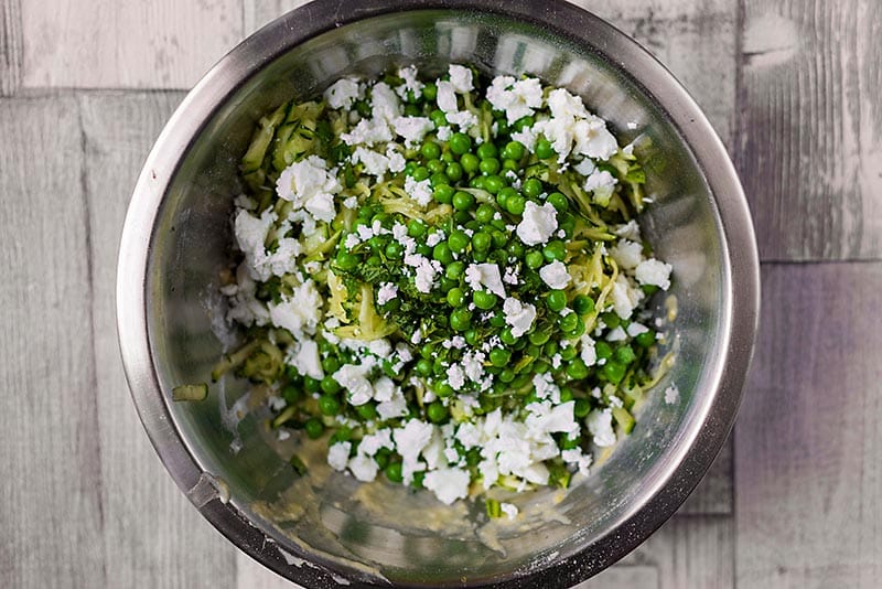 A mixing bowl containing grated zucchini, peas and feta.