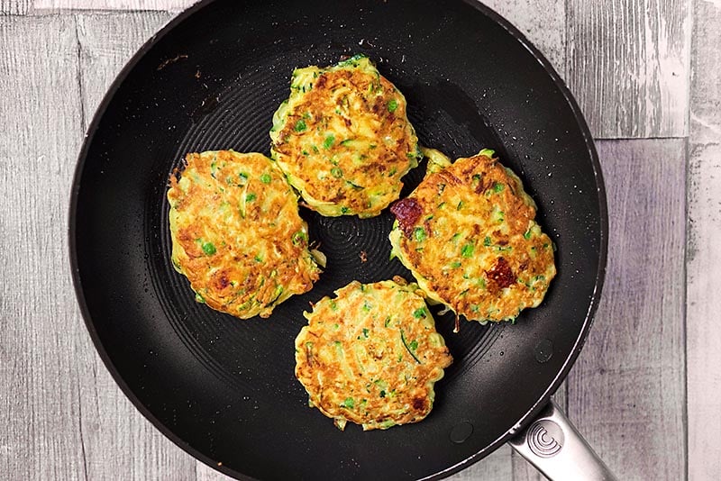 A frying pan with four courgette fritters cooking in it.