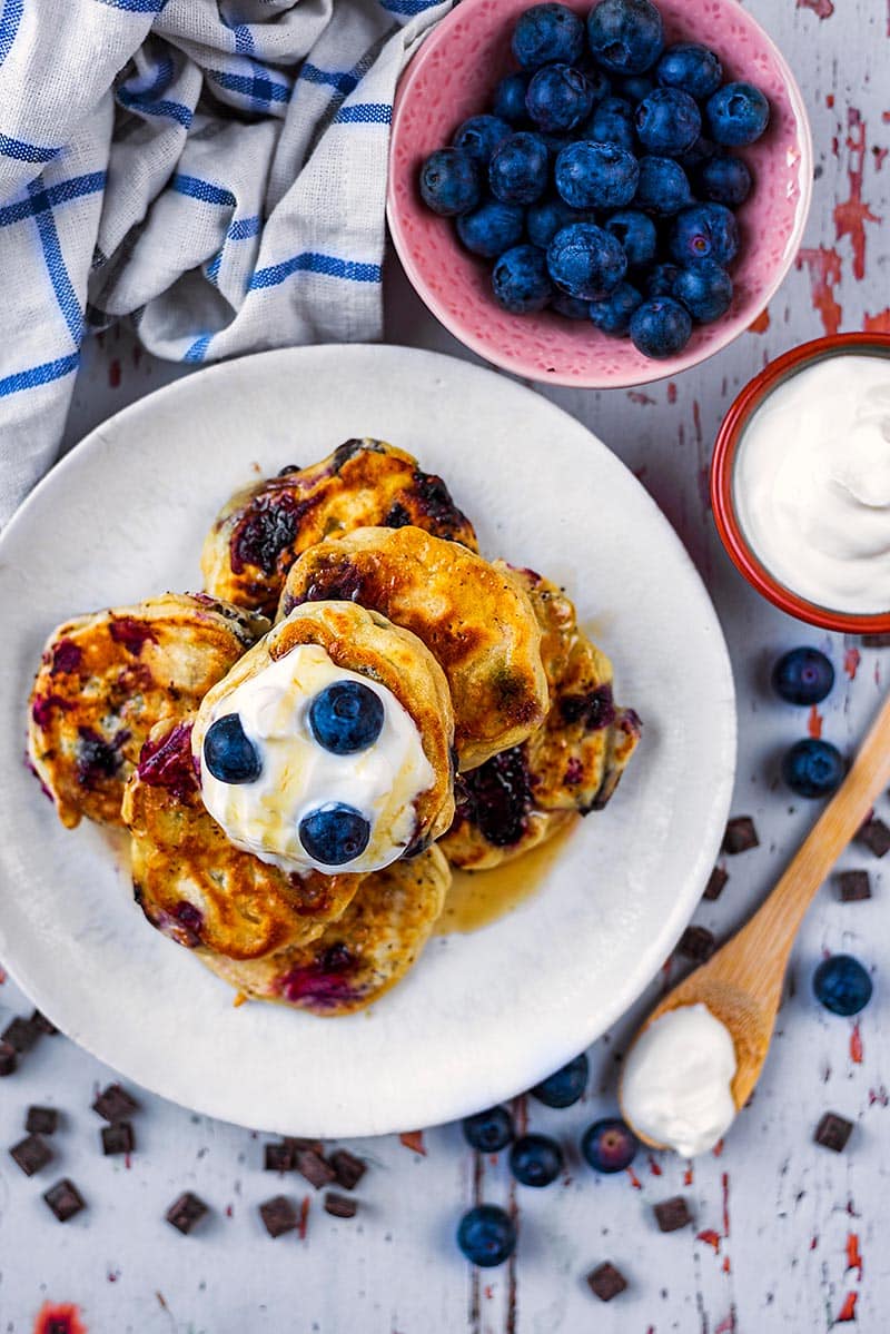 A plate of mini pancakes next to a bowl of blueberries and a bowl of yogurt.