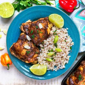 Two slow cooker jerk chicken thighs on a blue plate with rice and lime wedges.